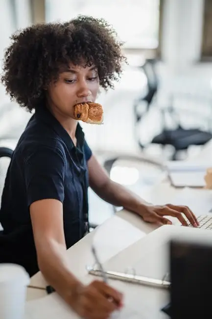 African American woman multitasking at office, eating pastry and working on laptop. Busy Professional Nutrition.